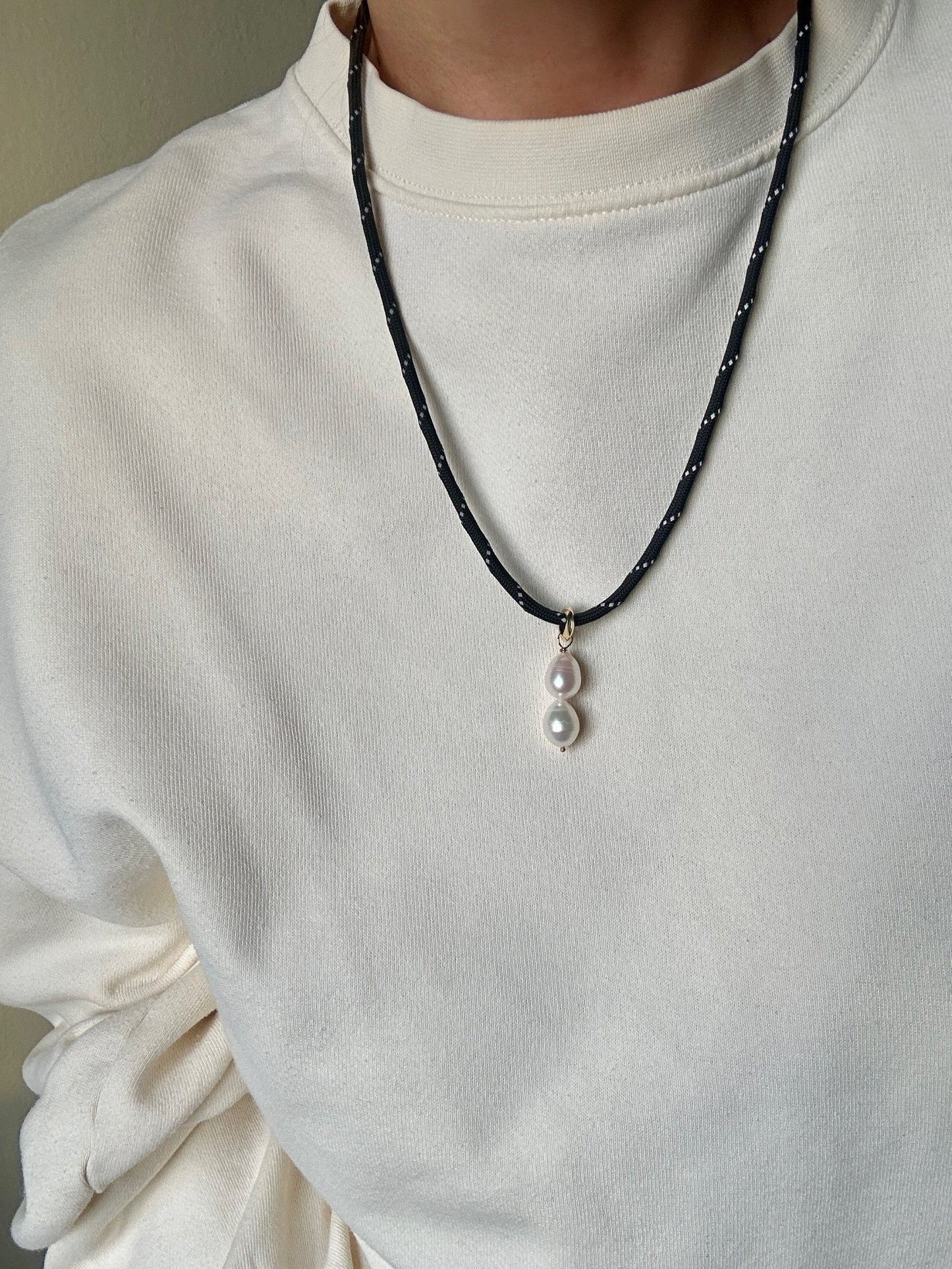 Person wearing a white shirt with a black paracord necklace featuring a pearl pendant.