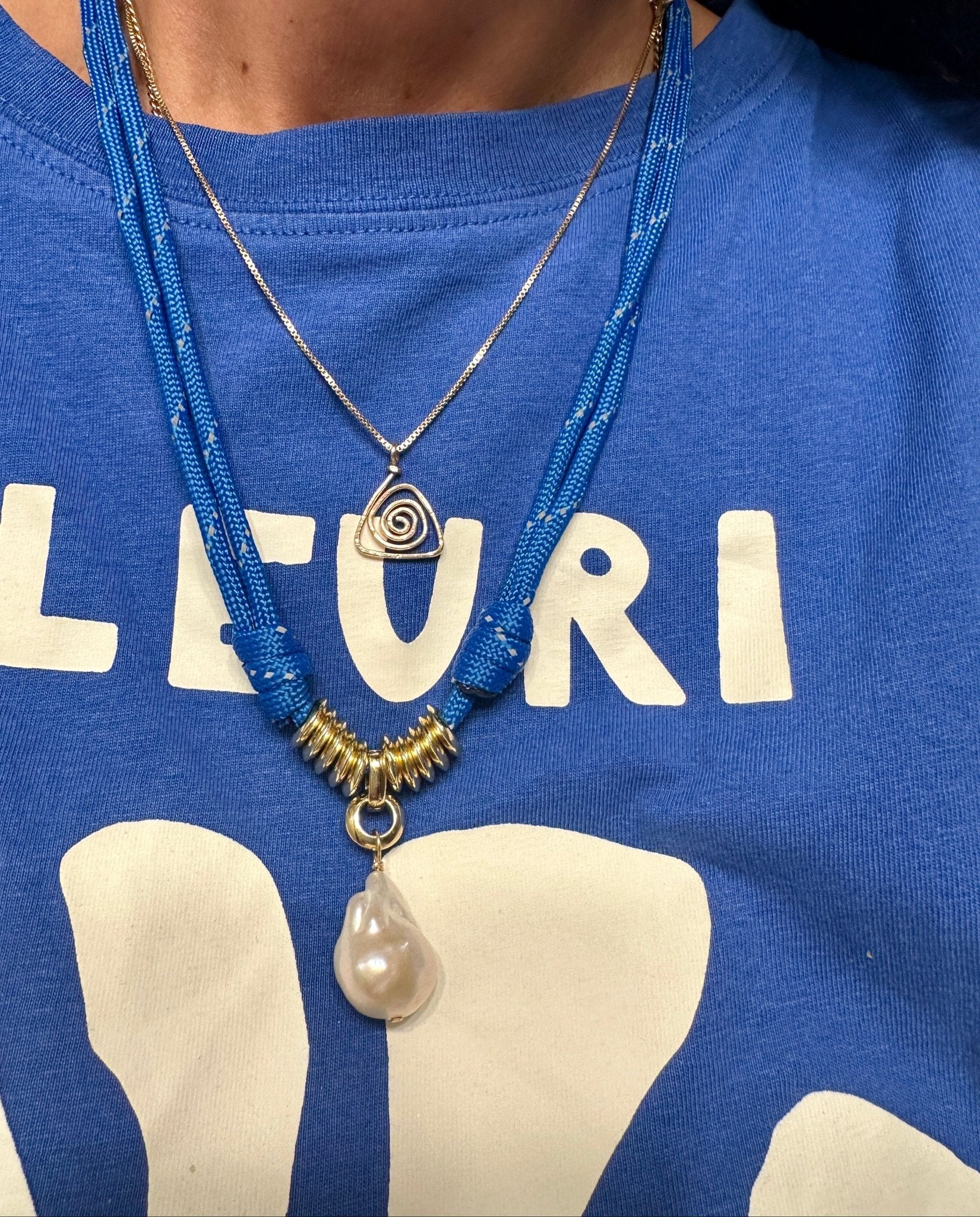 Woman wearing a gold triangle spiral necklace and a blue paracord and baroque pearl necklace on a blue shirt