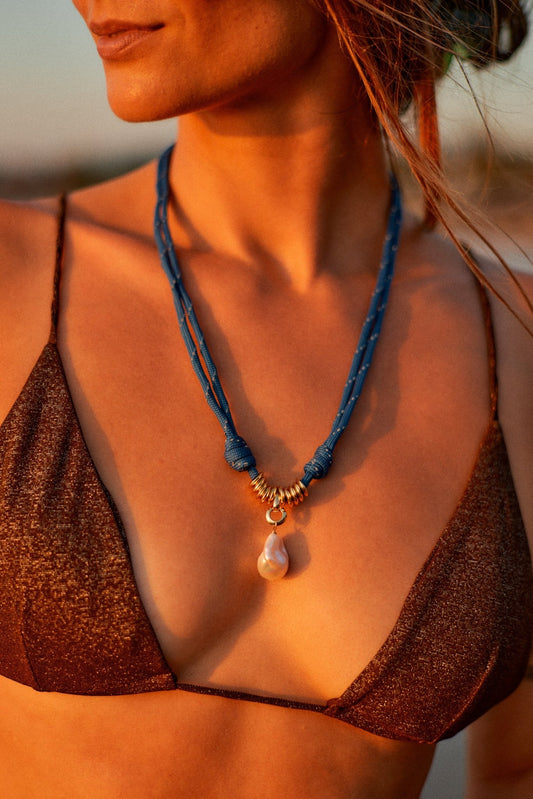 Woman wearing a blue paracord necklace with a pearl pendant, set against a blurred natural background