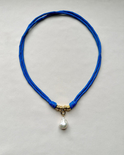 Flatlay of a blue paracord cord necklace with a baroque pearl pendant on a cream white background