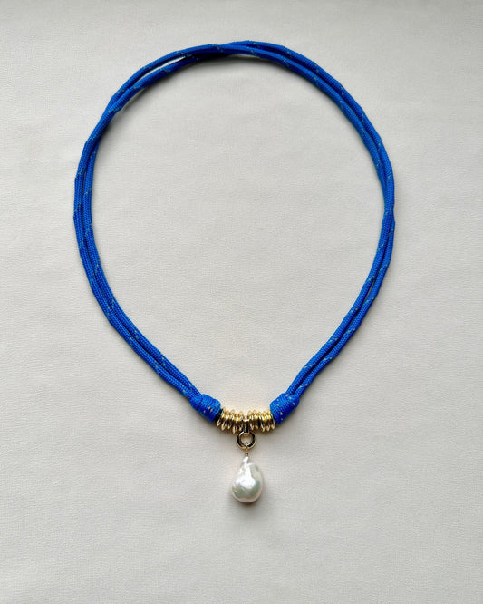 Flatlay of a blue paracord cord necklace with a baroque pearl pendant on a cream white background