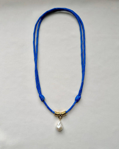 Flatlay of a blue paracord cord necklace with a baroque pearl pendant on a cream white background