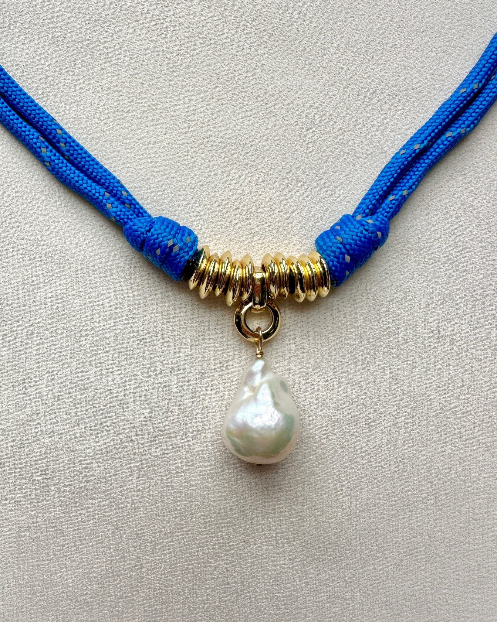 Close up of a necklace on a blue paracord cord with a baroque pearl pendant on a white background