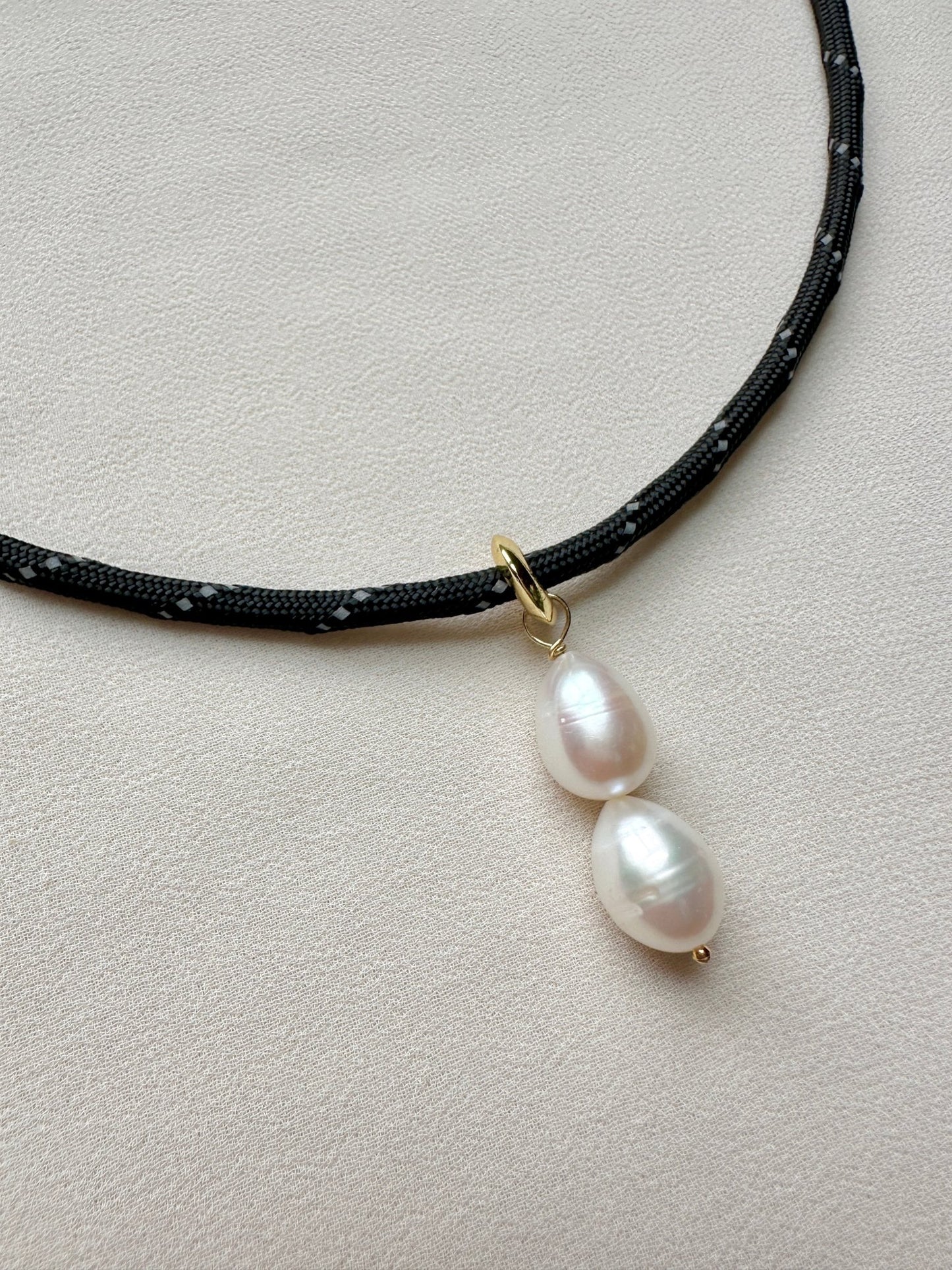 Close-up of the a black paracord necklace with freshwater pearl drop and gold plated bail on a fabric background.