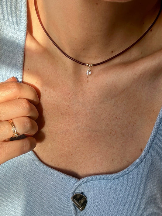 Woman wearing a minimalist burgundy leather cord choker necklace with a small teardrop gemstone pendant and gold accents. Styled with a light blue knit top and gold ring in natural sunlight.