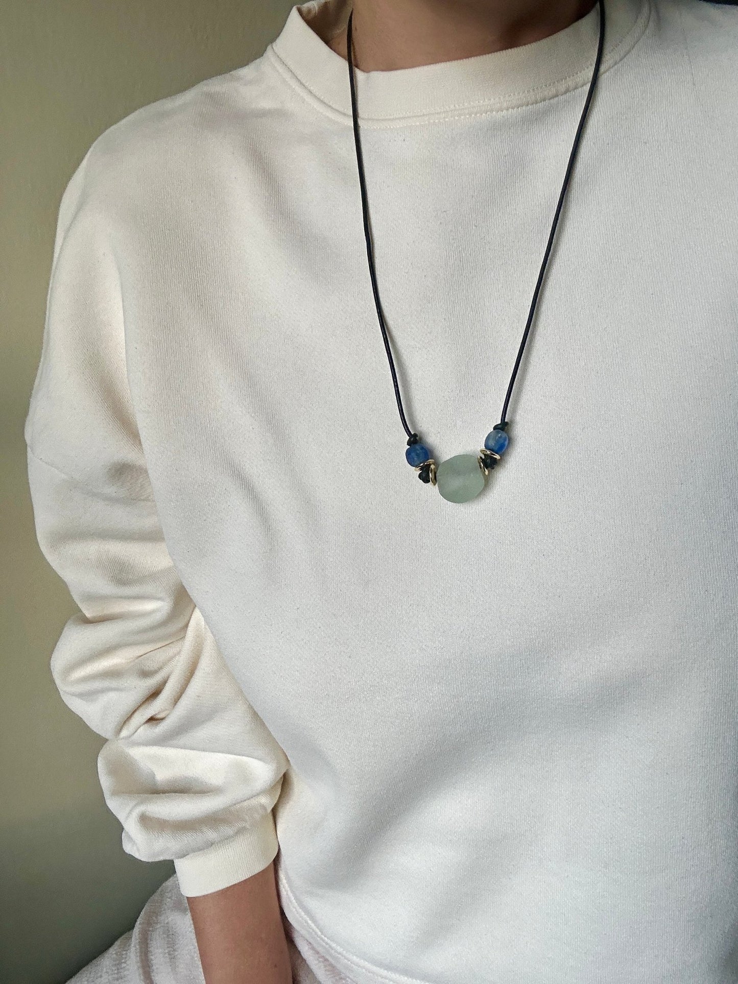 Person wearing a white sweater with a necklace featuring green and blue glass beads on a black leather cord.
