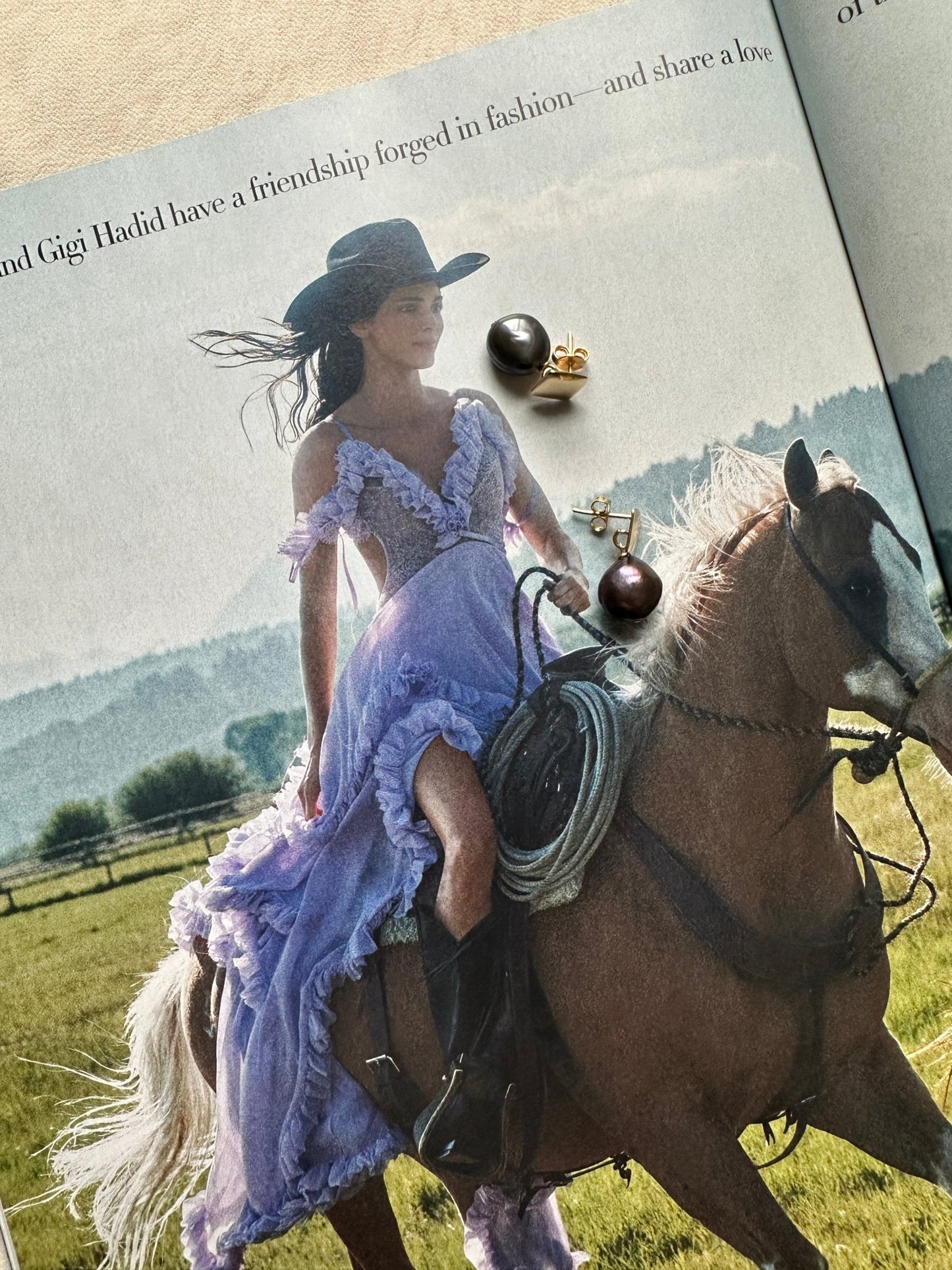 Magazine editorial of a woman in a purple dress riding a horse in a field with a flat lay of dark pearl stud earrings on top
