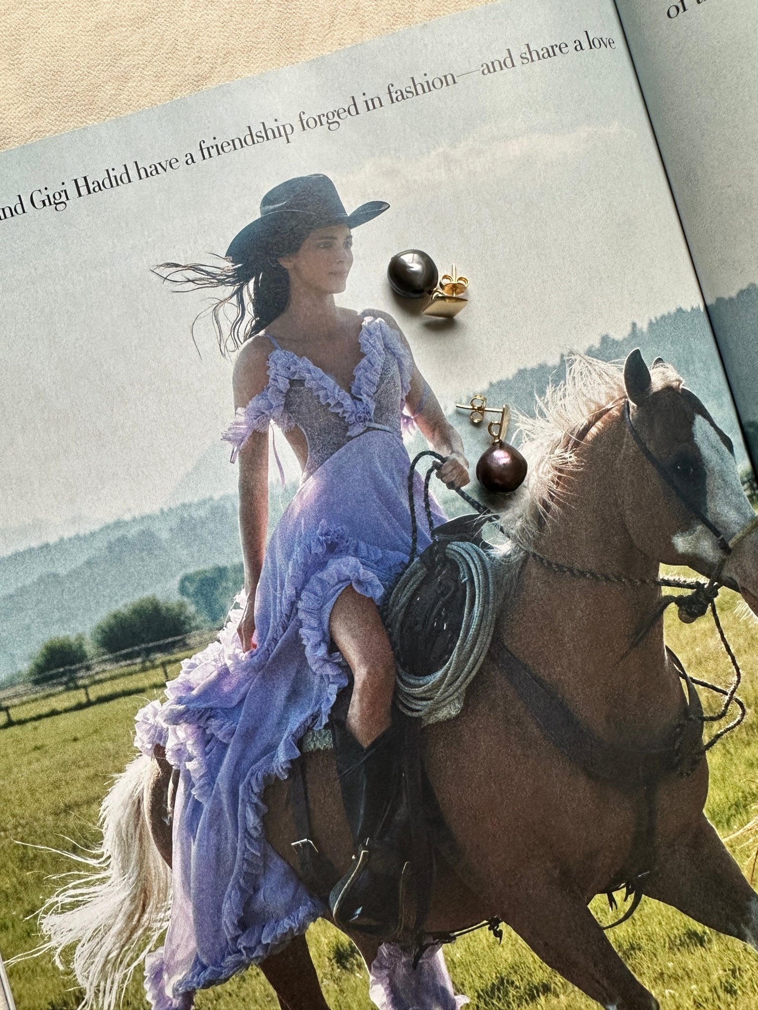 Magazine editorial of a woman in a purple dress riding a horse in a field with a flat lay of dark pearl stud earrings on top