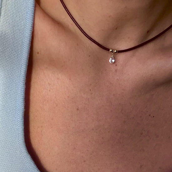 Close-up video of a minimalist burgundy leather cord choker necklace with a teardrop gemstone pendant and gold accents, worn with a light blue knit top.