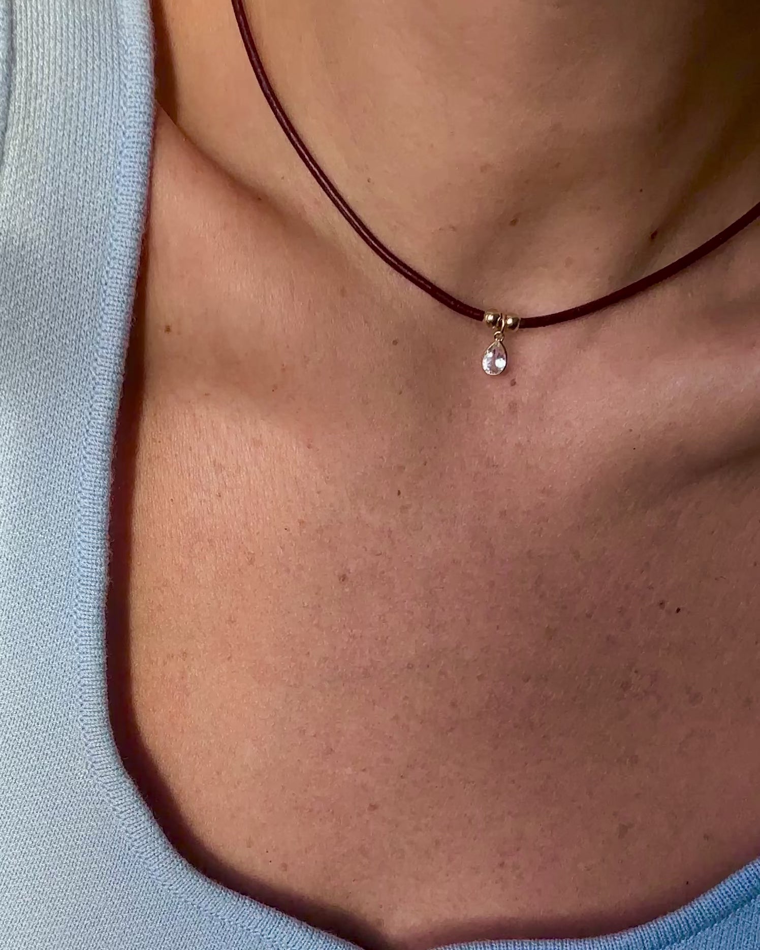 Close-up video of a minimalist burgundy leather cord choker necklace with a teardrop gemstone pendant and gold accents, worn with a light blue knit top.