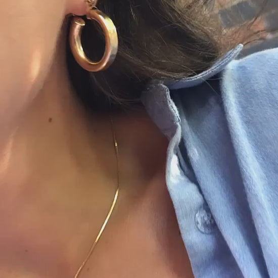 woman wearing thick gold hoop earrings and a thin glowy chain necklace paired with a buttoned blue shirt with flashes of sunshine and wind in her hair.