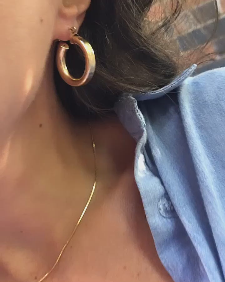 woman wearing thick gold hoop earrings and a thin glowy chain necklace paired with a buttoned blue shirt with flashes of sunshine and wind in her hair.